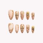 Theodora’s Golden Cross: nude and gold-tone almond press-on nails with ornate cross motifs and textured metallic accents, set arranged on a pale background.