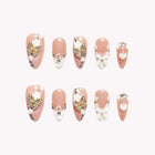 Psyche’s Winged Ribbon: nude almond press-on nails with white flowers and bows, butterfly-like motifs and sparkling gold-tone accents, set arranged on a pale background.