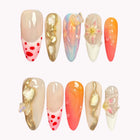 Hesperia’s Golden Charm: long almond press-on nails with orange gradients, red dots, iridescent swirls and gold-tone raised accents, set arranged on a pale background.