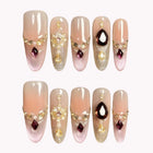 Guinevere’s Garnet Tear: nude almond press-on nails with dark teardrop gems and gold-tone decorations, set arranged on a pale background.