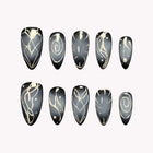 Nyx’s Chrome Sigil: dark blue-gray almond press-on nails with silver-tone spirals, lattice lines and starbursts, set arranged on a pale background.