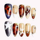 Stellar Amber Drift almond nails with amber tortoiseshell patterns, black and nude panels, white stars and metallic studs.