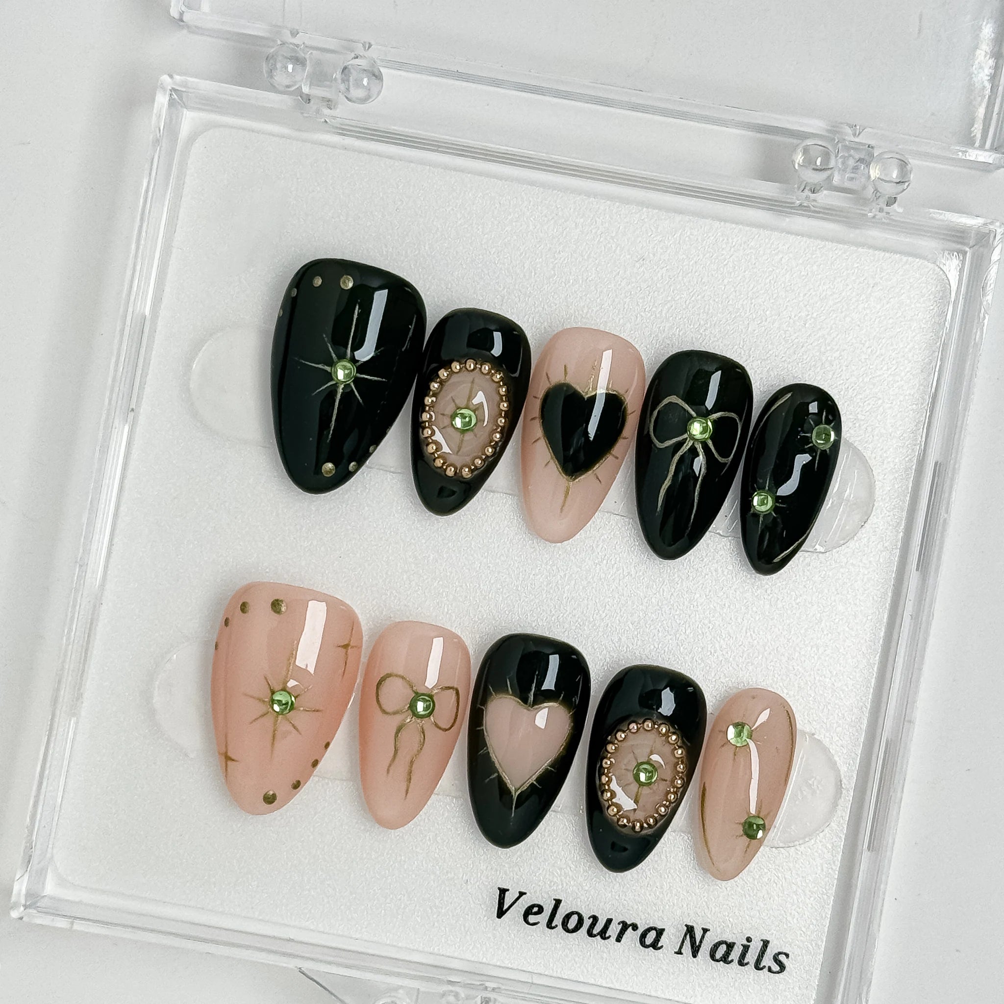 Black and nude heart-accent nails with green gems arranged inside a clear Veloura Nails display case.