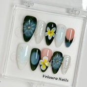 Deep green and milky white floral nails arranged inside a clear Veloura Nails display case.