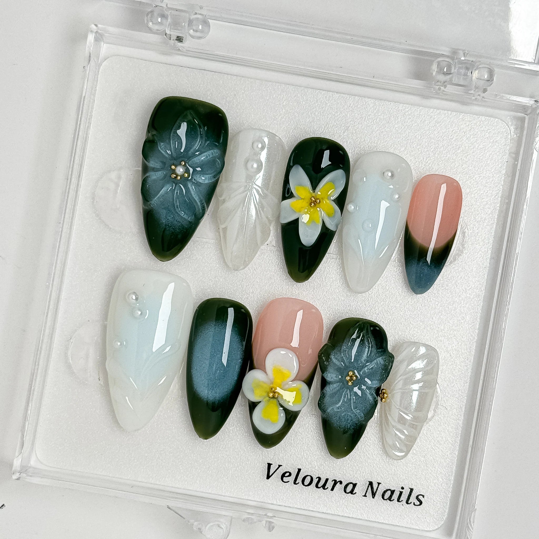 Deep green and milky white floral nails arranged inside a clear Veloura Nails display case.