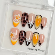 Vintage Havana Bloom: yellow, nude and brown almond press-on nails with spotted patterns, white flowers and starfish accents, displayed in a clear Veloura Nails case.