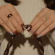 Helios’ Amber Amulet: brown, taupe and silver-tone almond press-on nails with oval metallic accents and fine starbursts, shown on hands holding a bag handle.