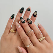 Overlapping hands with gold-colored rings wearing black and nude nails with hearts and green gem accents.