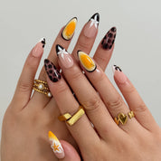 Vintage Havana Bloom: yellow, nude and brown almond press-on nails with spotted patterns, white flowers and starfish accents, shown on crossed fingers with gold-tone rings.