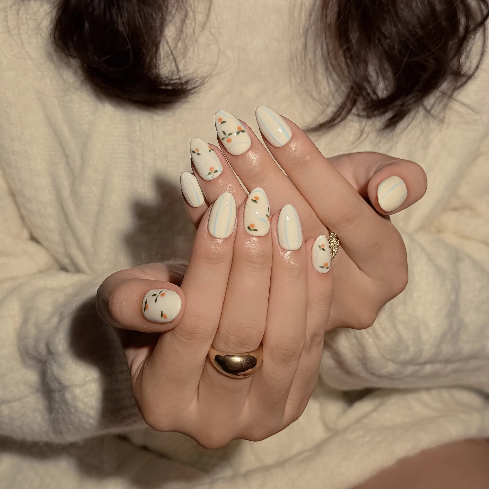 Pomona’s Azure Grove: cream press-on nails with pale blue stripes and tiny orange fruit motifs, worn with cream knit sleeves.