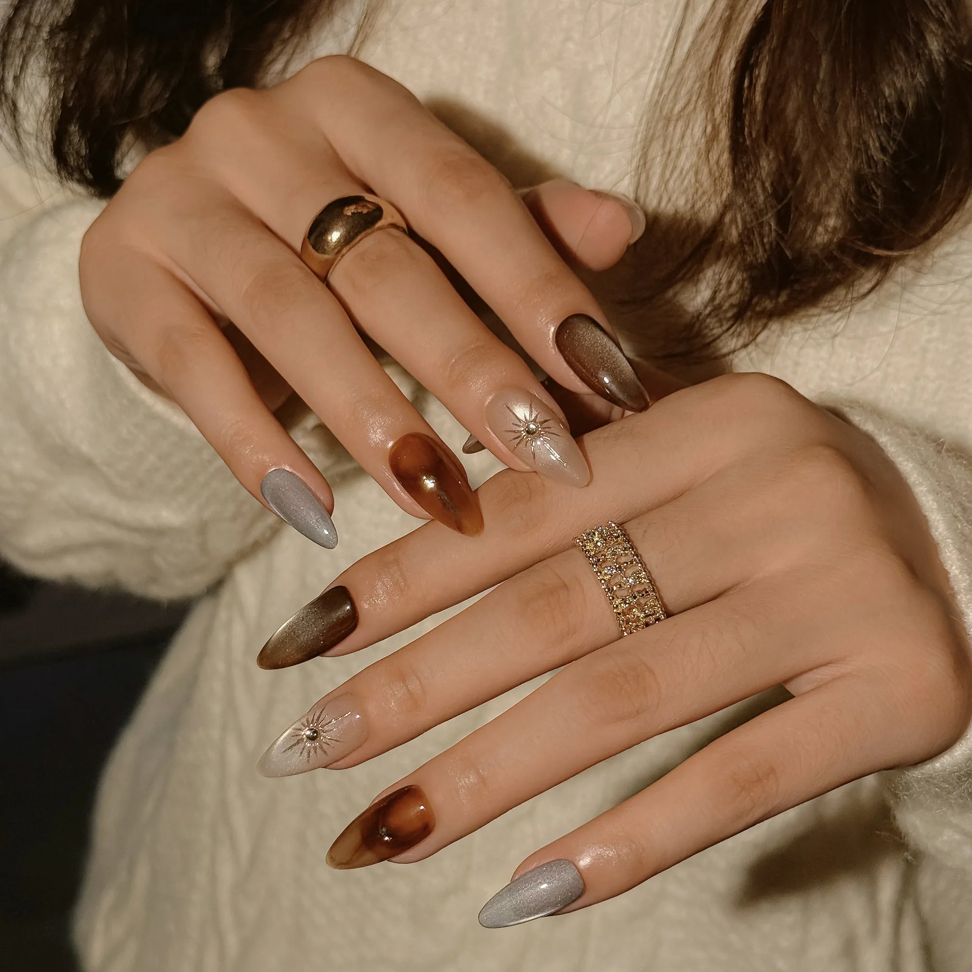 Helios’ Amber Amulet: brown, taupe and silver-tone almond press-on nails with oval metallic accents and fine starbursts, shown on overlapping hands against a cream sweater.
