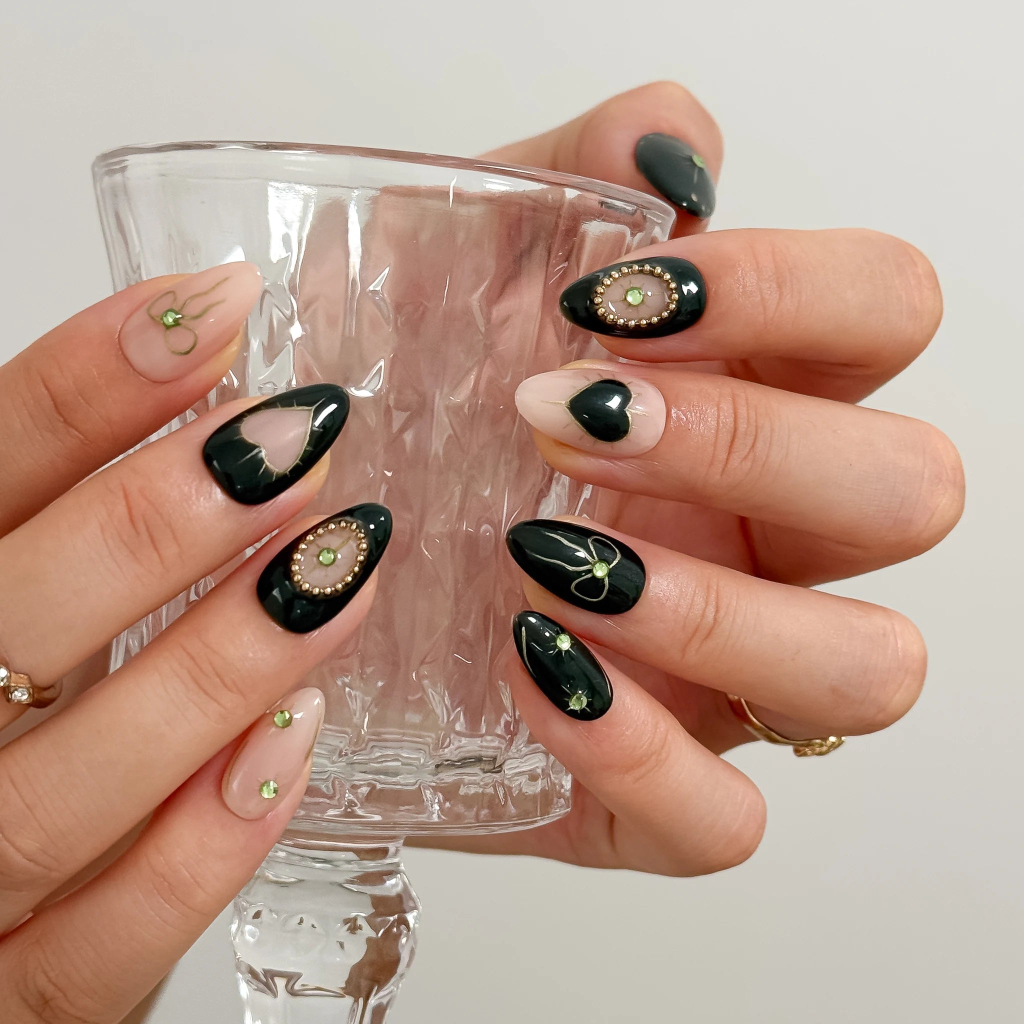 Hands holding a clear stemmed glass, showing black heart motifs and oval green decorations on nude nails.