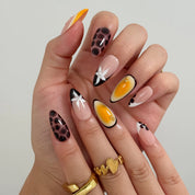 Vintage Havana Bloom: yellow, nude and brown almond press-on nails with spotted patterns, white flowers and starfish accents, close-up of curled fingers.