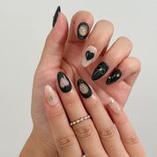 Curled and resting hand poses showing black and nude almond nails with gold-colored detailing.