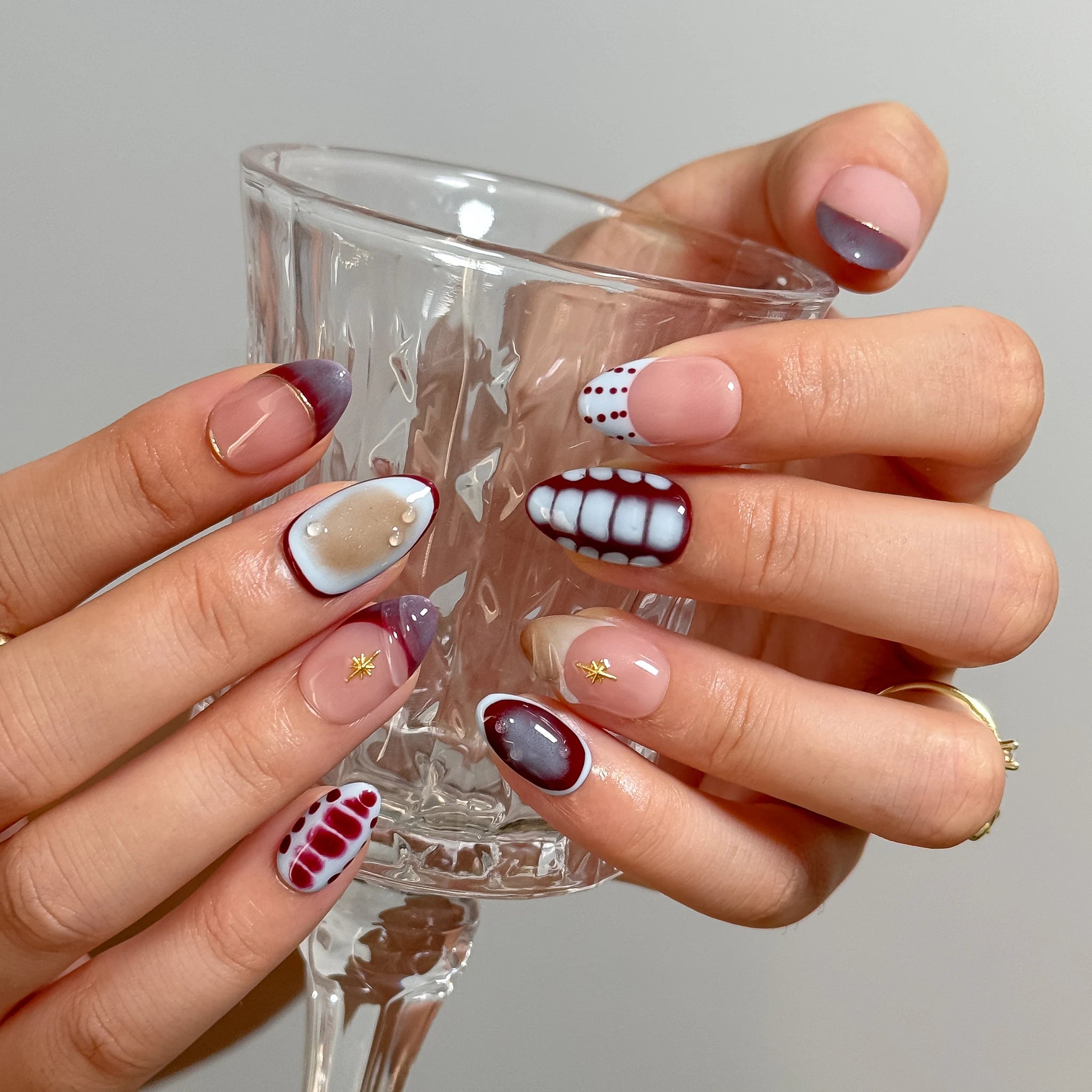 Chimera’s Amber Mosaic press-on nails holding a clear glass, showing burgundy and nude pattern details.