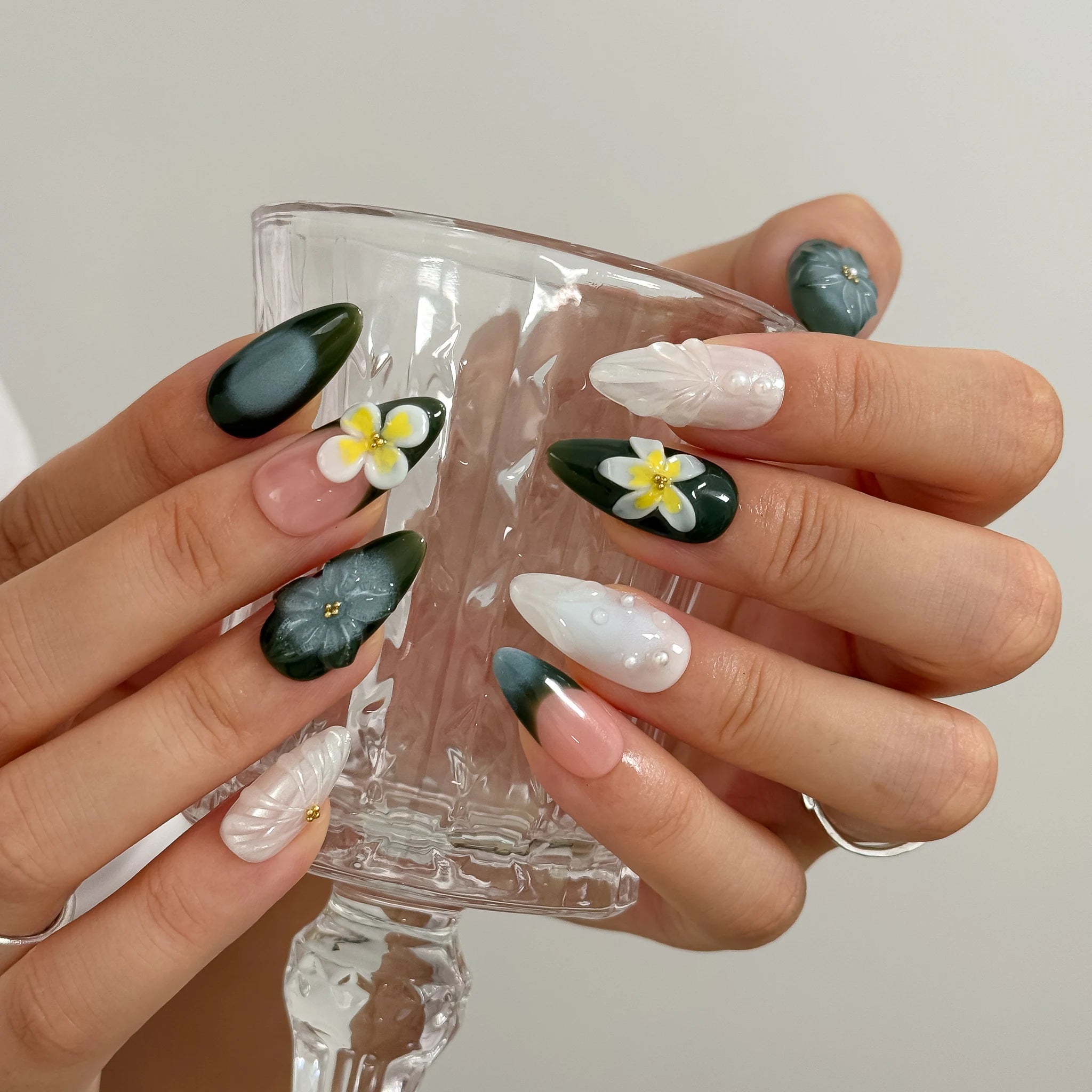 Hands holding a clear stemmed glass, showing sculpted flowers on deep green and milky white nails.
