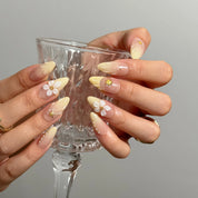 Nereid's Radiance press-on nails holding a clear glass, showing yellow ombré and floral details.