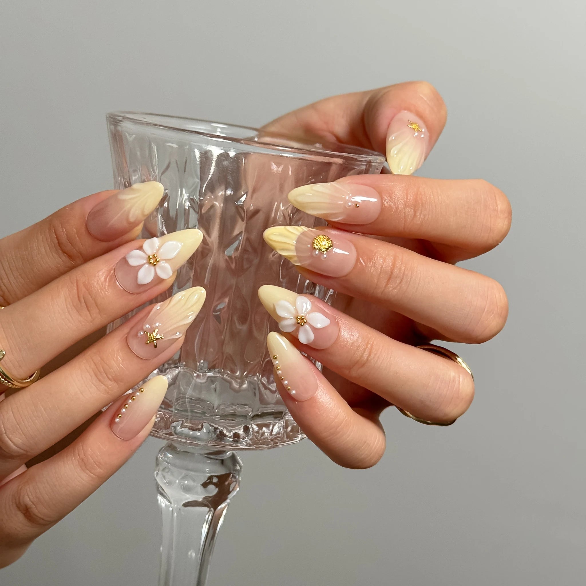 Nereid's Radiance press-on nails holding a clear glass, showing yellow ombré and floral details.