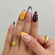 Vintage Havana Bloom: yellow, nude and brown almond press-on nails with spotted patterns, white flowers and starfish accents, modeled on raised fingers against gray.