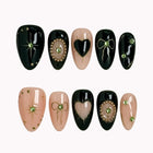 Astra's Ebony Locket black and nude almond nails with hearts, fine gold-colored bows and green oval gem accents.
