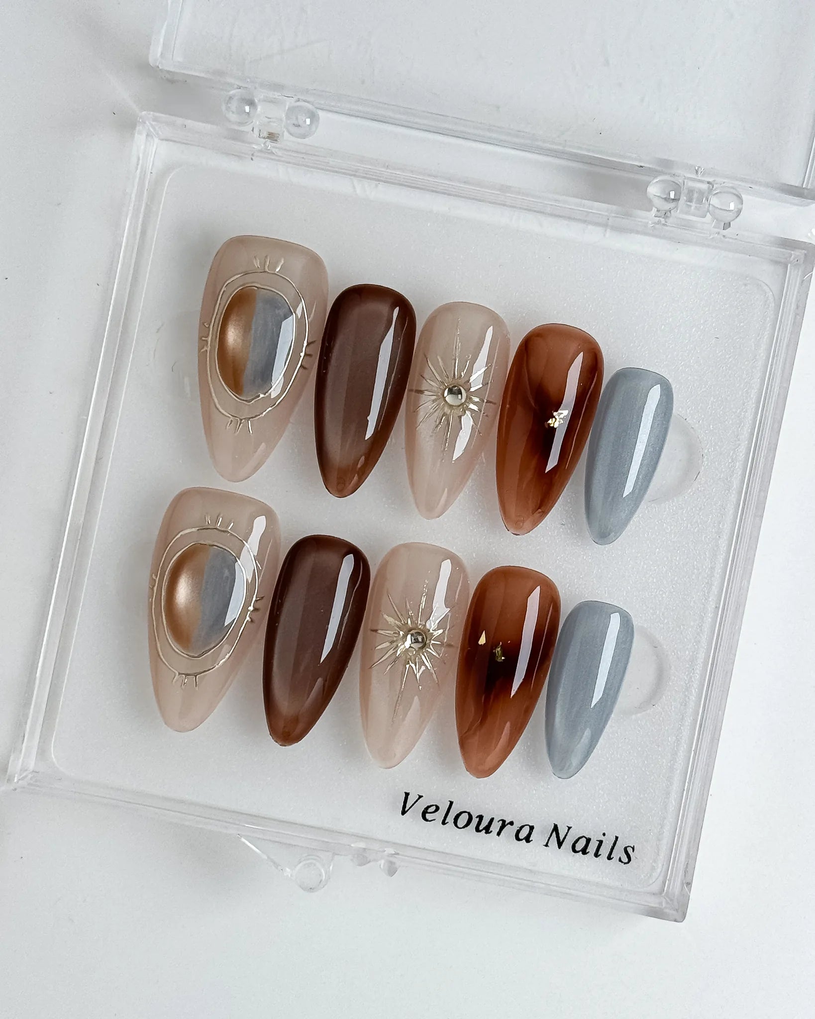 Helios’ Amber Amulet: brown, taupe and silver-tone almond press-on nails with oval metallic accents and fine starbursts, displayed in a clear Veloura Nails case.