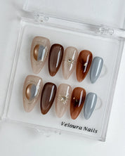 Helios’ Amber Amulet: brown, taupe and silver-tone almond press-on nails with oval metallic accents and fine starbursts, displayed in a clear Veloura Nails case.
