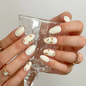 Pomona’s Azure Grove: cream press-on nails with pale blue stripes and tiny orange fruit motifs, worn while holding a small glass.