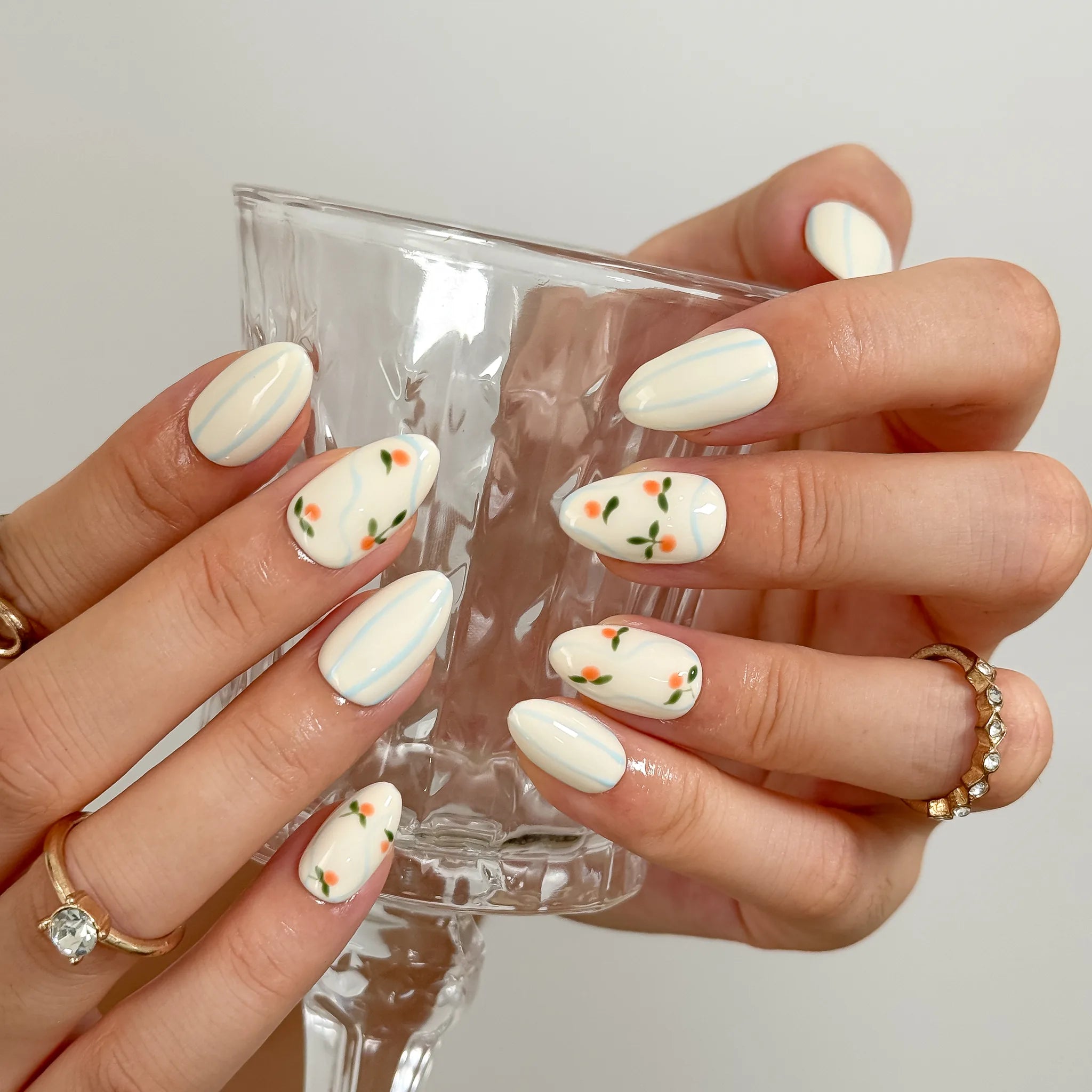 Pomona’s Azure Grove: cream press-on nails with pale blue stripes and tiny orange fruit motifs, worn while holding a small glass.