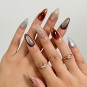 Helios’ Amber Amulet: brown, taupe and silver-tone almond press-on nails with oval metallic accents and fine starbursts, shown on crossed fingers.