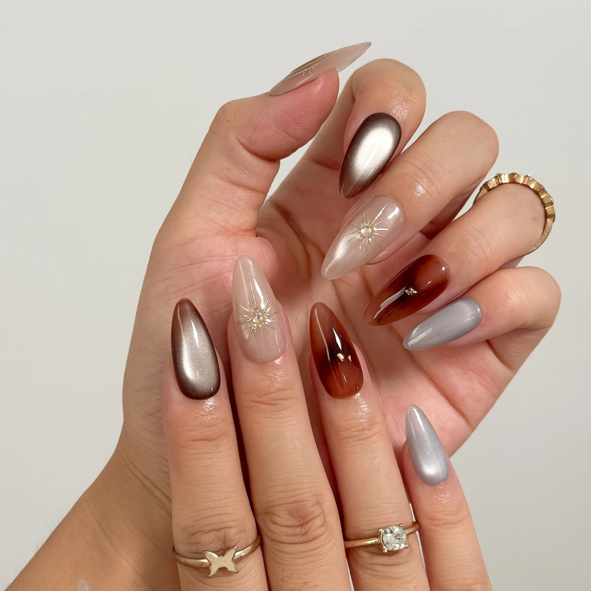Helios’ Amber Amulet: brown, taupe and silver-tone almond press-on nails with oval metallic accents and fine starbursts, close-up of curled fingers.