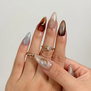 Helios’ Amber Amulet: brown, taupe and silver-tone almond press-on nails with oval metallic accents and fine starbursts, modeled on raised fingers with rings.