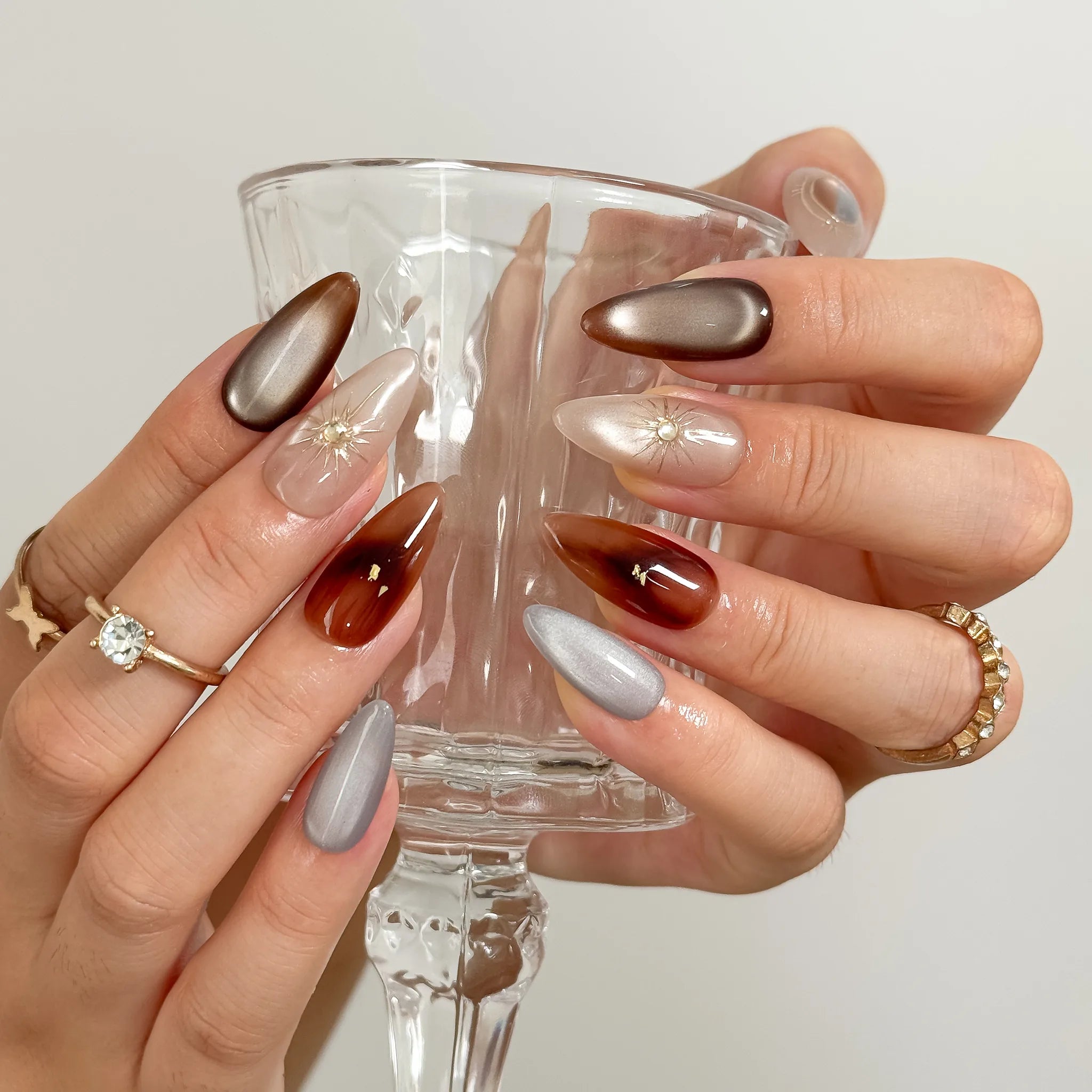 Helios’ Amber Amulet: brown, taupe and silver-tone almond press-on nails with oval metallic accents and fine starbursts, worn while holding a stemmed glass.