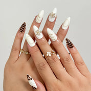 Bastet’s Desert Flower: white and brown almond press-on nails with spotted patterns, metallic outlines and star accents, shown on crossed fingers with rings.