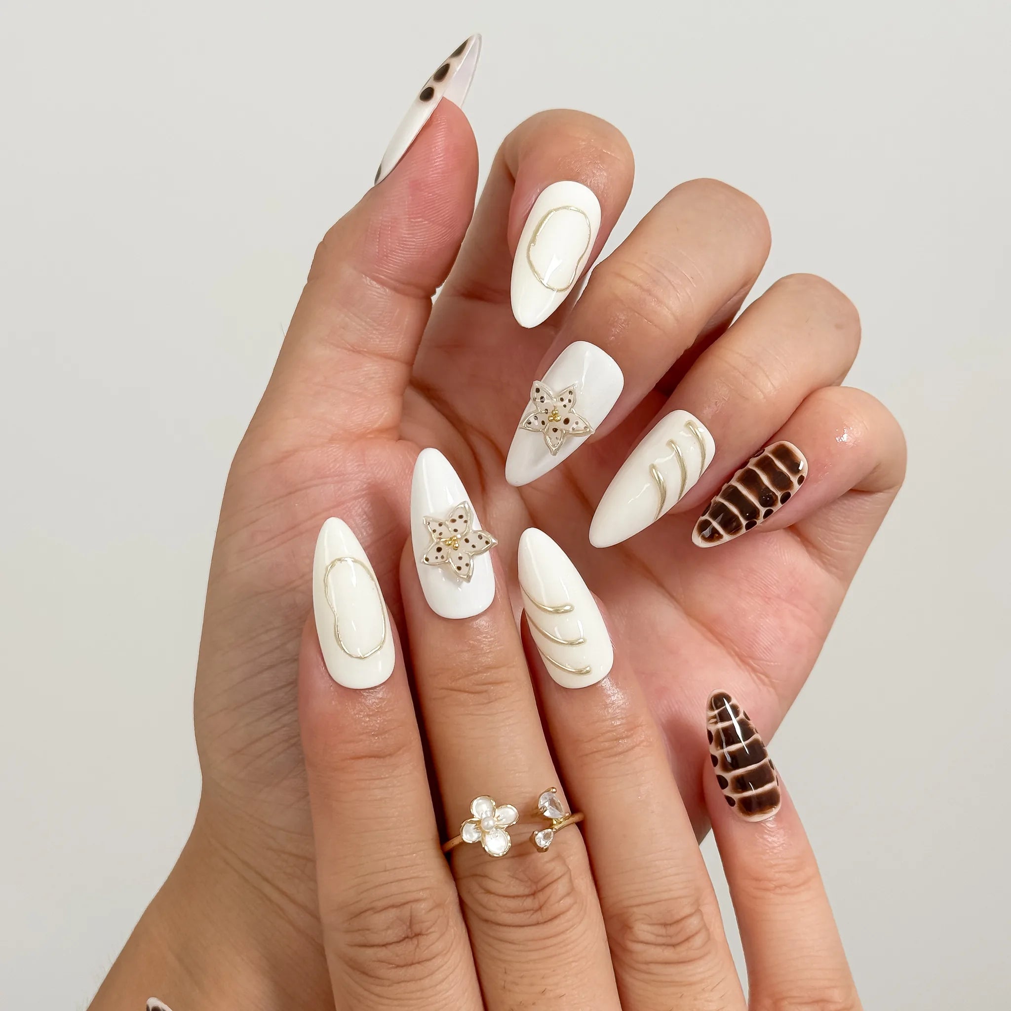 Bastet’s Desert Flower: white and brown almond press-on nails with spotted patterns, metallic outlines and star accents, close-up of curled fingers.