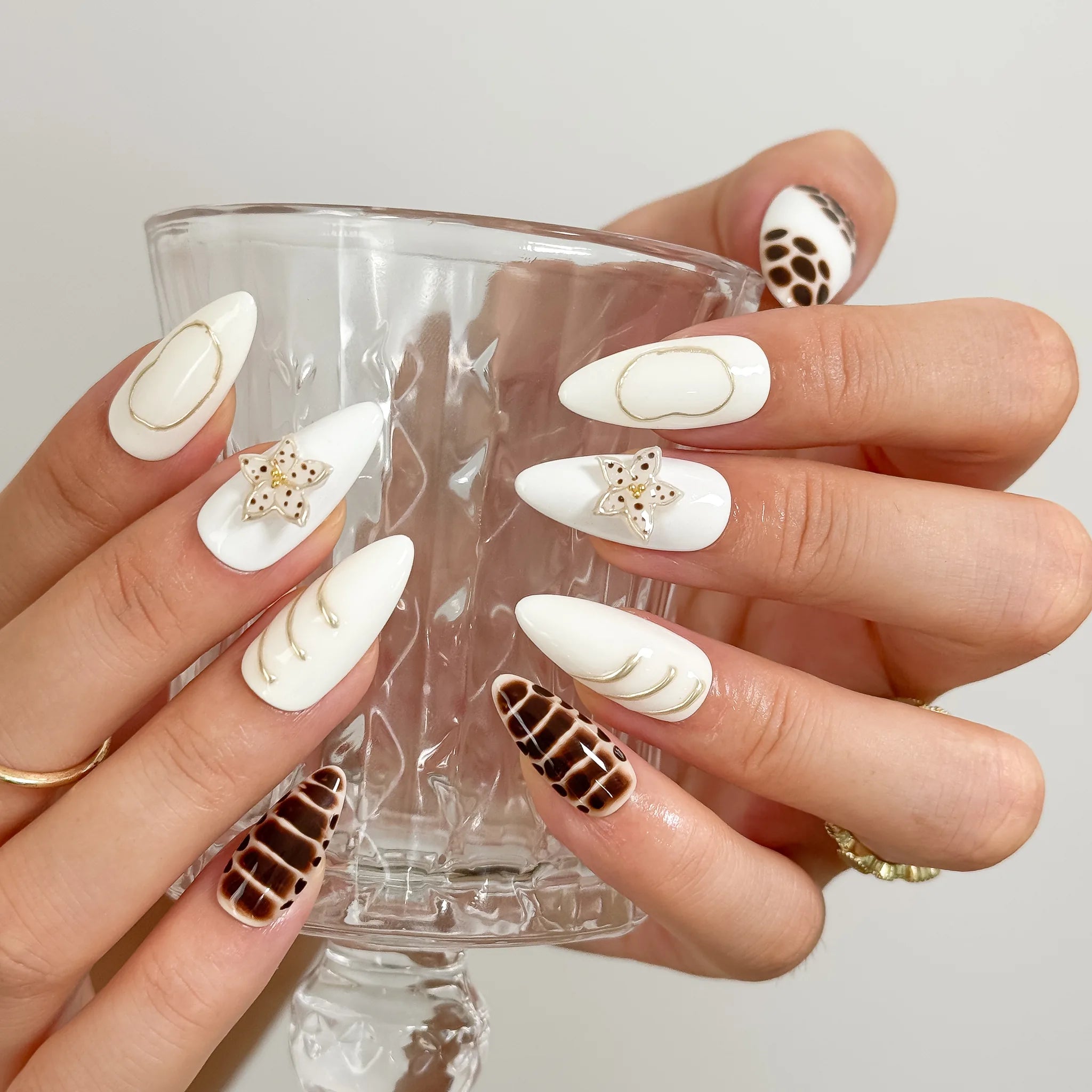 Bastet’s Desert Flower: white and brown almond press-on nails with spotted patterns, metallic outlines and star accents, worn while holding a stemmed glass.