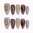 Helios’ Amber Amulet: brown, taupe and silver-tone almond press-on nails with oval metallic accents and fine starbursts, set arranged on a pale background.
