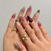 Crimson Rosary Bloom long almond press-on nails with pink aura tones, gold beads, and crystal cross accents shown on hand