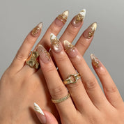 Gilded Magnolia Veil long almond press-on nails with raised gold flowers and white French tips shown on hand