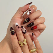 Cherry Noir Petals long almond press-on nails with black cherry tones, white dots, sculpted pink flowers, and gold details shown on hand