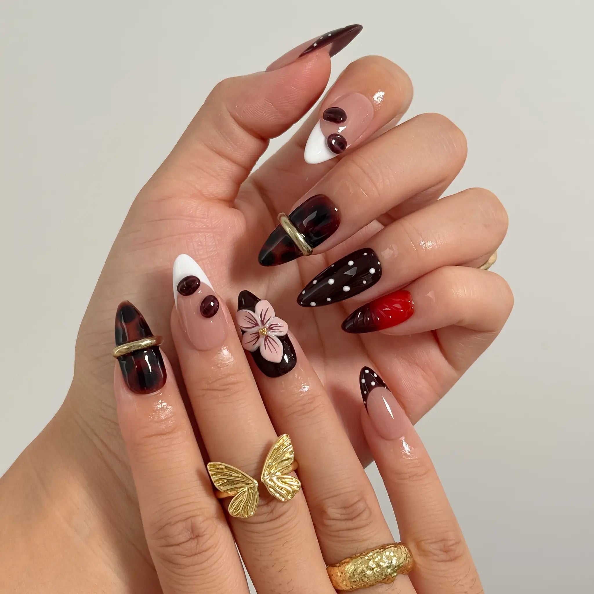 Cherry Noir Petals long almond press-on nails with black cherry tones, white dots, sculpted pink flowers, and gold details shown on hand