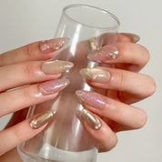 Daphne’s Golden Laurel: nude and gold-toned almond press-on nails with iridescent flakes and raised metallic textures, worn while holding a tall glass.