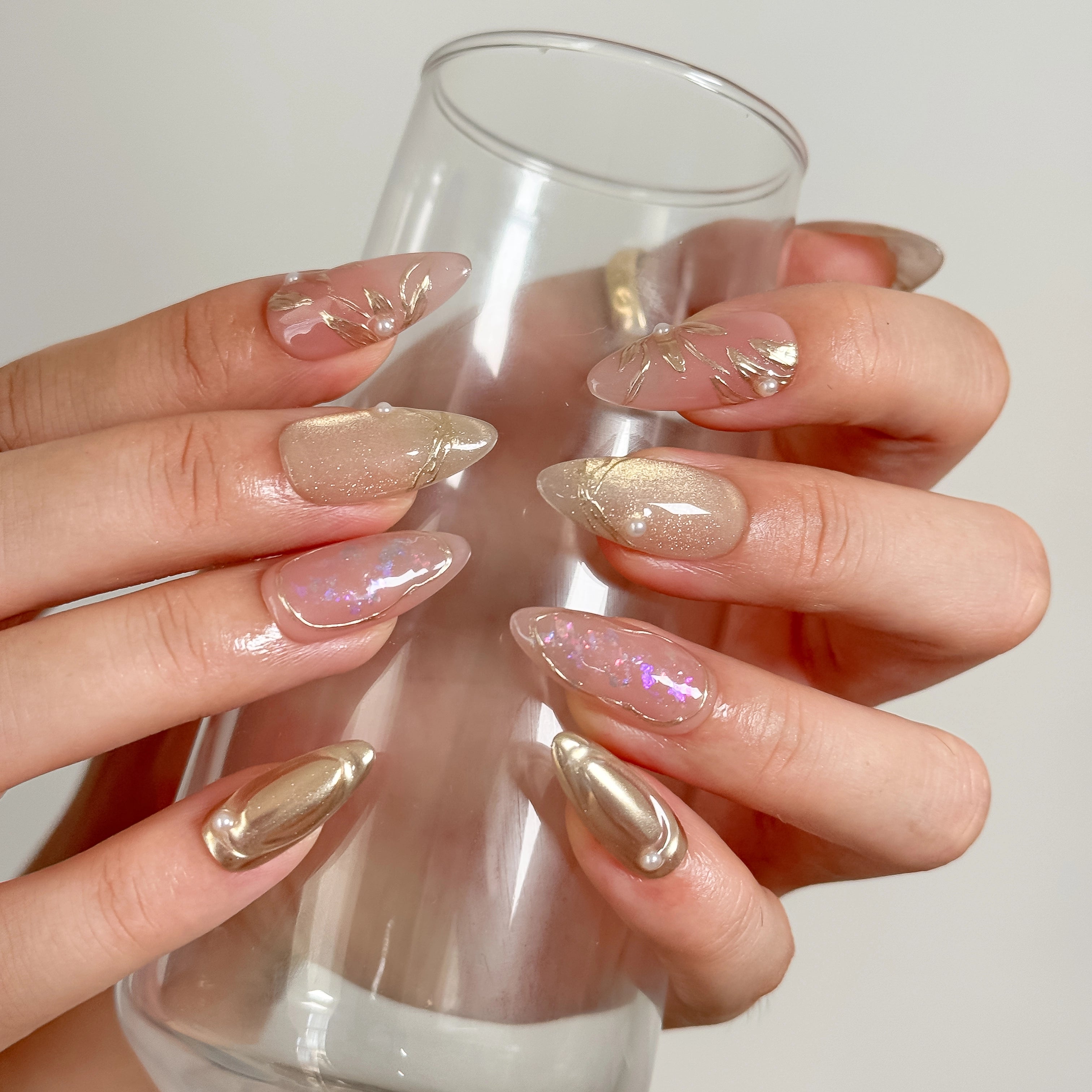 Daphne’s Golden Laurel: nude and gold-toned almond press-on nails with iridescent flakes and raised metallic textures, worn while holding a tall glass.