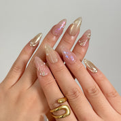 Daphne’s Golden Laurel: nude and gold-toned almond press-on nails with iridescent flakes and raised metallic textures, shown on raised hands.