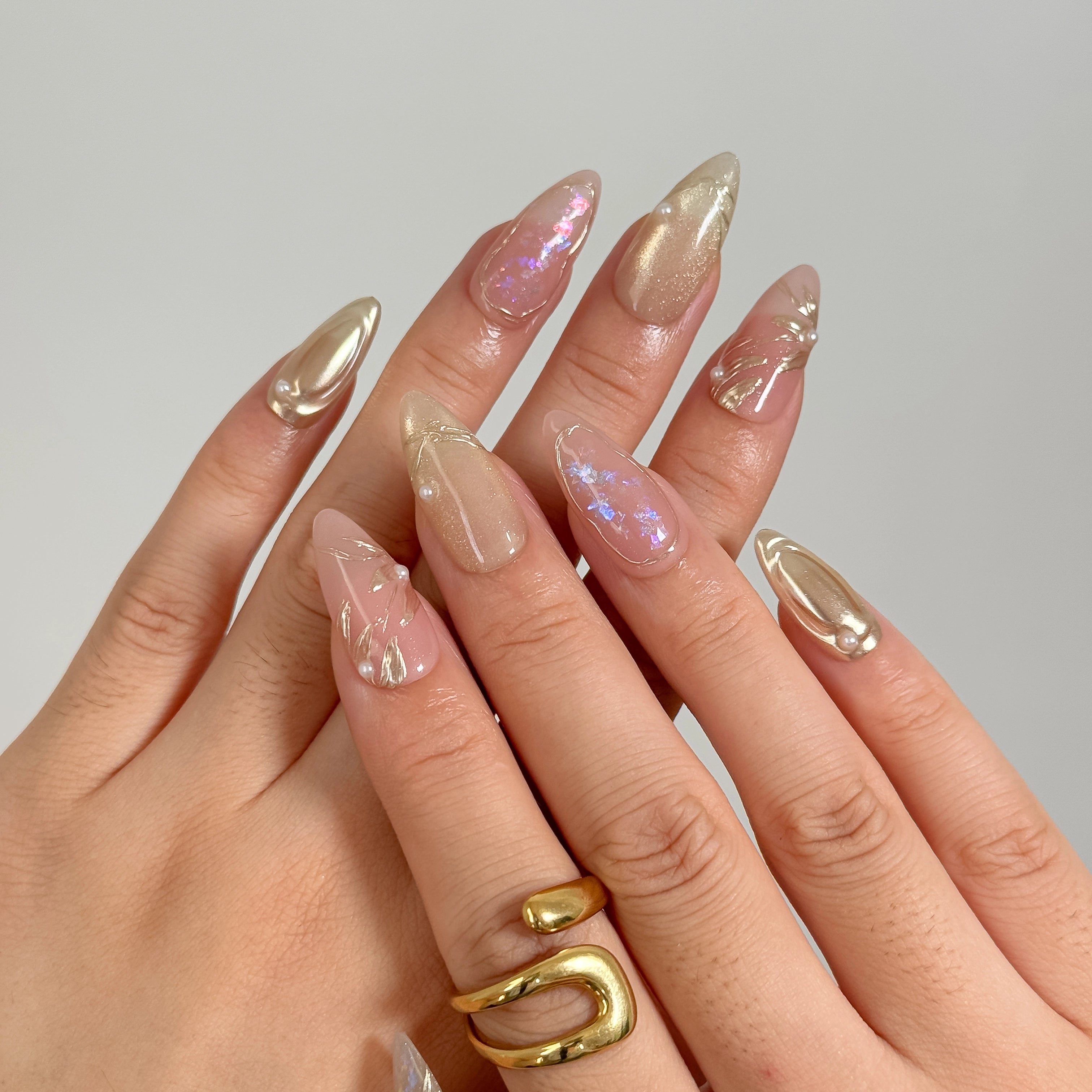 Daphne’s Golden Laurel: nude and gold-toned almond press-on nails with iridescent flakes and raised metallic textures, shown on raised hands.