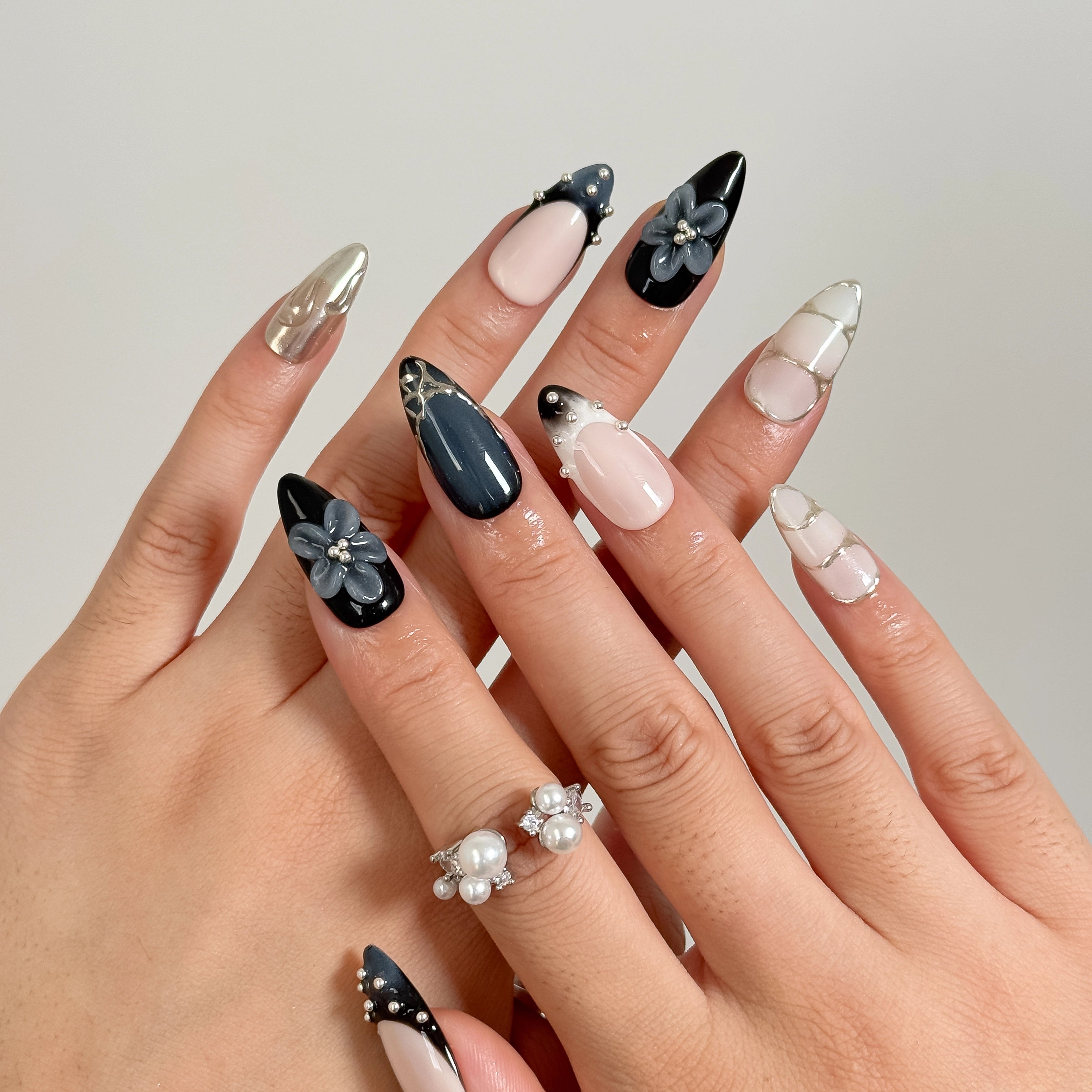 Midnight Alloy Bloom: black, nude and silver-tone almond press-on nails with raised gray flowers and studded tips, shown on crossed fingers against gray.