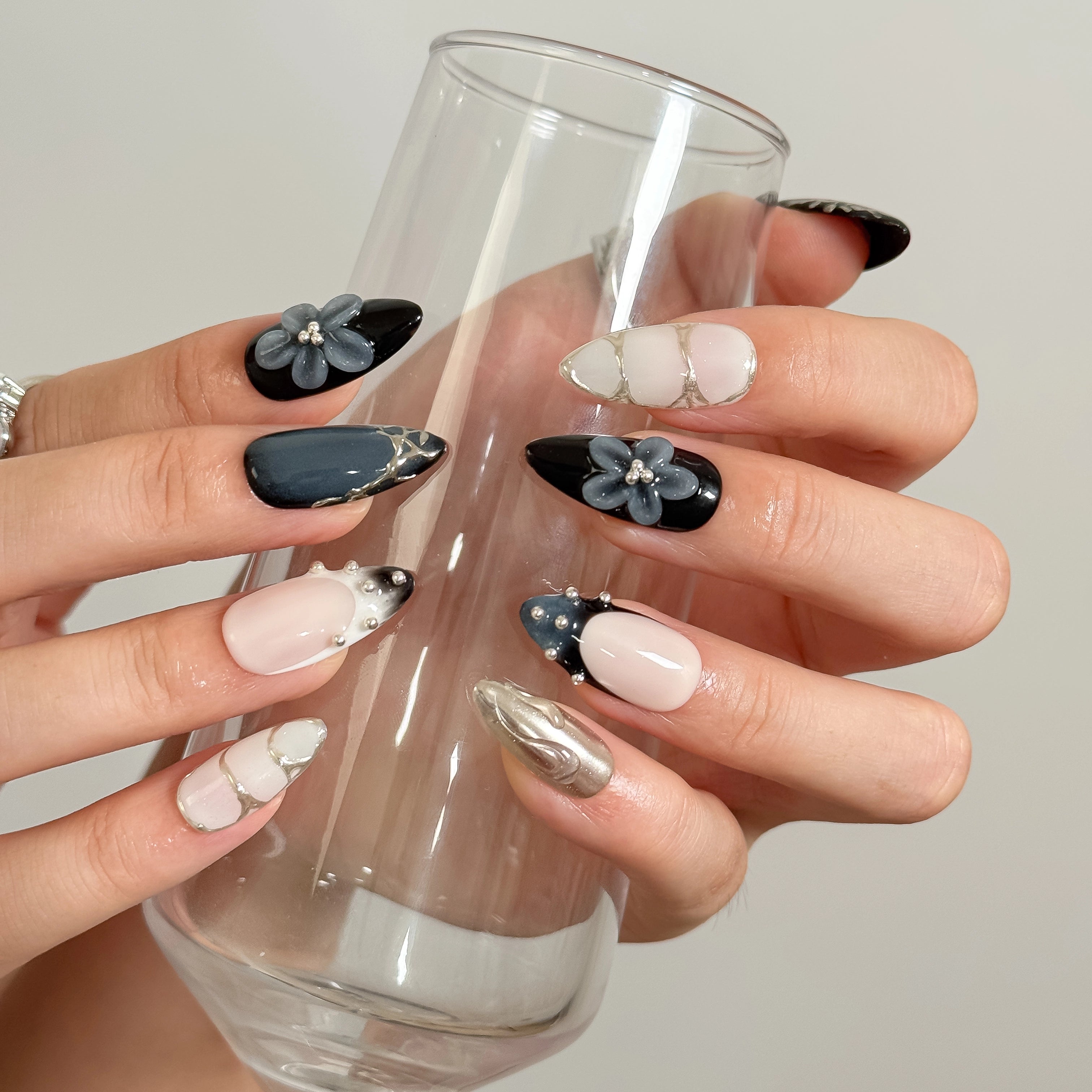 Midnight Alloy Bloom: black, nude and silver-tone almond press-on nails with raised gray flowers and studded tips, worn while holding a tall clear glass.