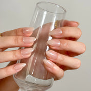 Champagne Silk Aura: glossy nude almond press-on nails with a pearly champagne sheen, worn while holding a tall glass.