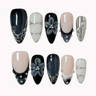 Midnight Alloy Bloom: black, nude and silver-tone almond press-on nails with raised gray flowers and studded tips, set arranged on a pale background.